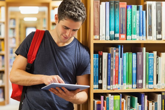 Student leaning against bookshelf holding a tablet pc at the library